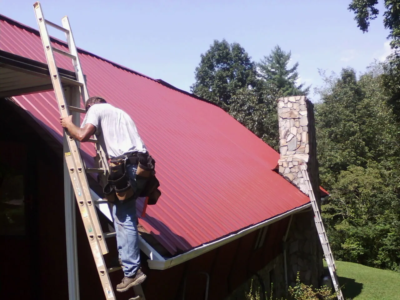 Roofer on a ladder beside a red metal roof and stone chimney — Soffit & Fascia Repair by Roofing Brothers in South Lyme